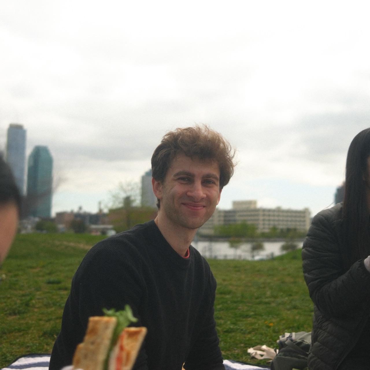 Aymeric smiling at a picnic in a park, a city skyline and river behind him