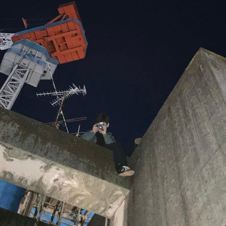 Aymeric perched on a concrete ledge at night, pointing a camera down at the viewer